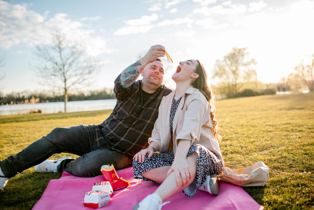 Man comically feeds fiancée McDonald's chips in open field at sunset.