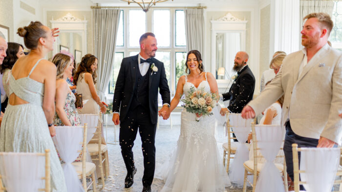 A bride and groom walk hand in hand down the aisle after their wedding ceremony at Horwood House. The bride wears a white lace gown and holds a bouquet of pale pink roses and greenery. The groom, in a classic black tuxedo, grins as he holds her hand. Guests throw confetti in celebration, smiling and clapping as they surround the couple. The elegant interior features light-colored curtains and decorative elements, creating a joyful atmosphere.