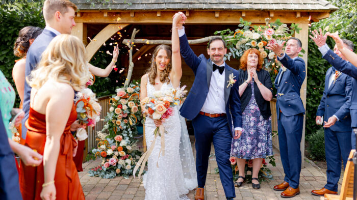 Happy couple celebrates after outdoor wedding ceremony under a wooden arbor adorned with flowers. Guests clap and toss petals, creating a joyful atmosphere.